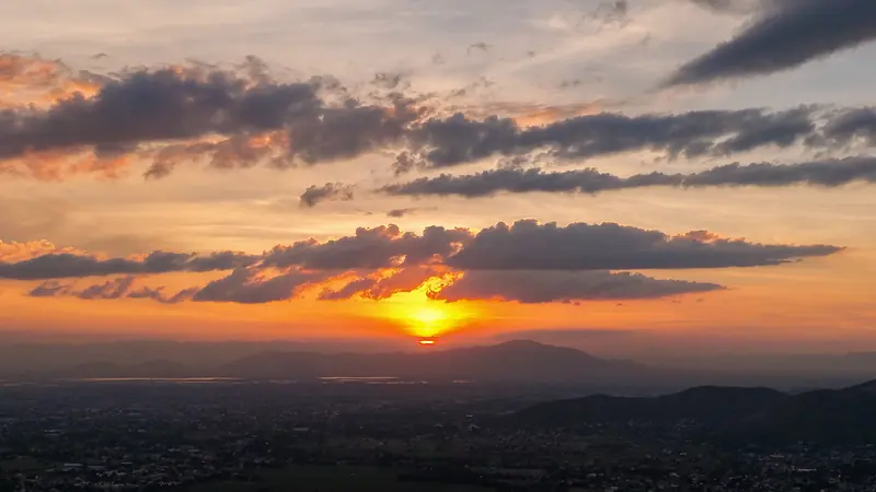 Atardecer visto desde el aire sobre un valle con montañas, con nubes oscuras y el sol naranja sobre el horizonte